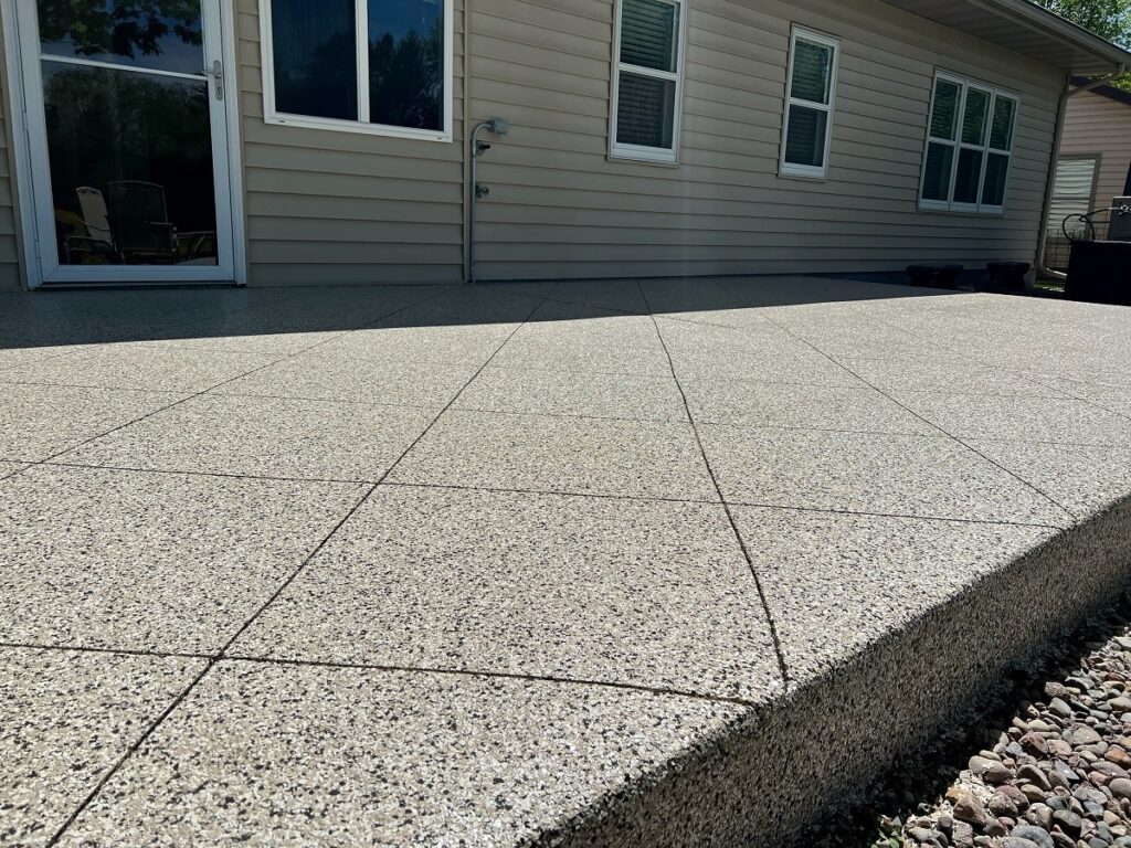 A large, textured patio in a backyard, adjacent to a beige house with windows and a sliding glass door, under clear sunlight.