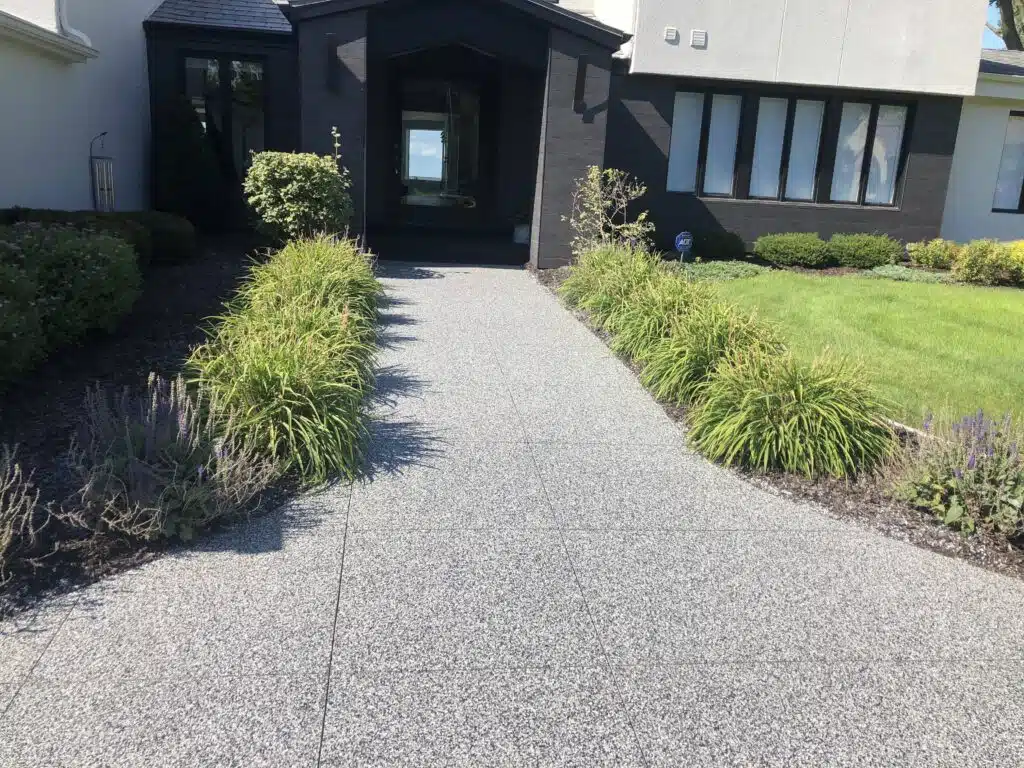 A modern house entrance with a grey path, surrounded by lush green plants and well-manicured grass, leading to a dark door.