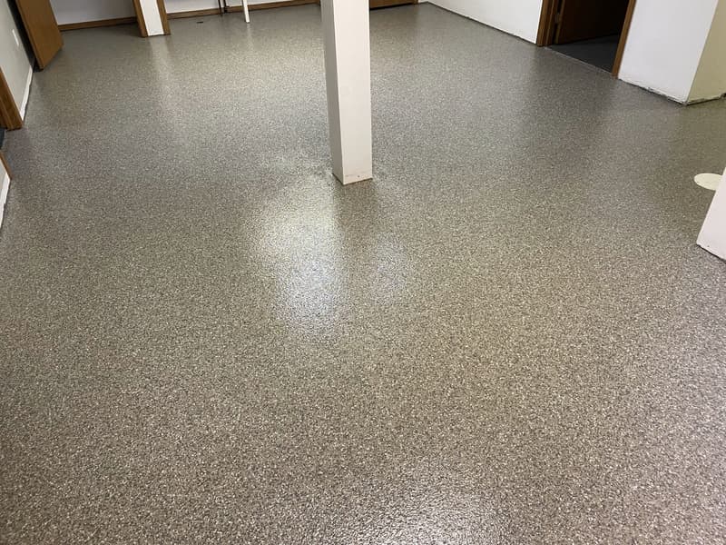 A vacant room with speckled gray flooring, white walls, and a central support column. No furniture or distinctive features present.