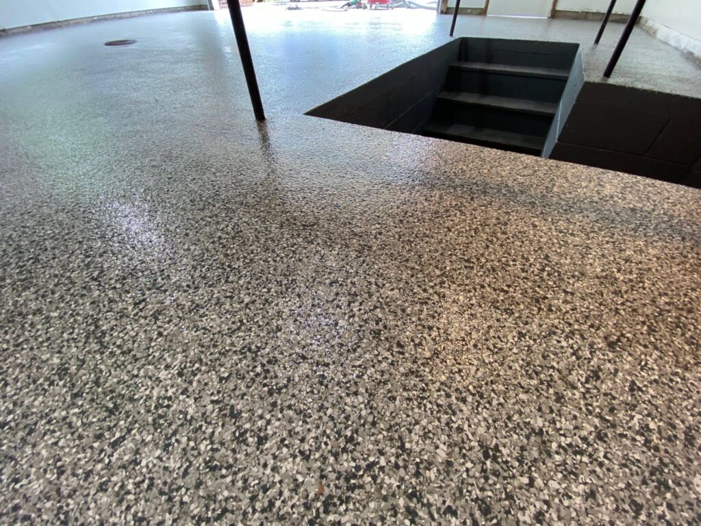 Epoxy-coated concrete floor with a speckled gray surface, leading to a staircase descending into a basement. Bright lighting enhances the glossy finish.