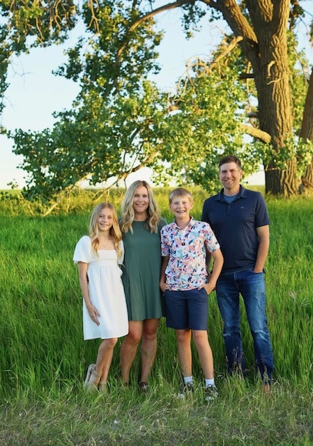 A family of four poses outdoors on green grass, with trees in the background. The group includes two children and two adults.
