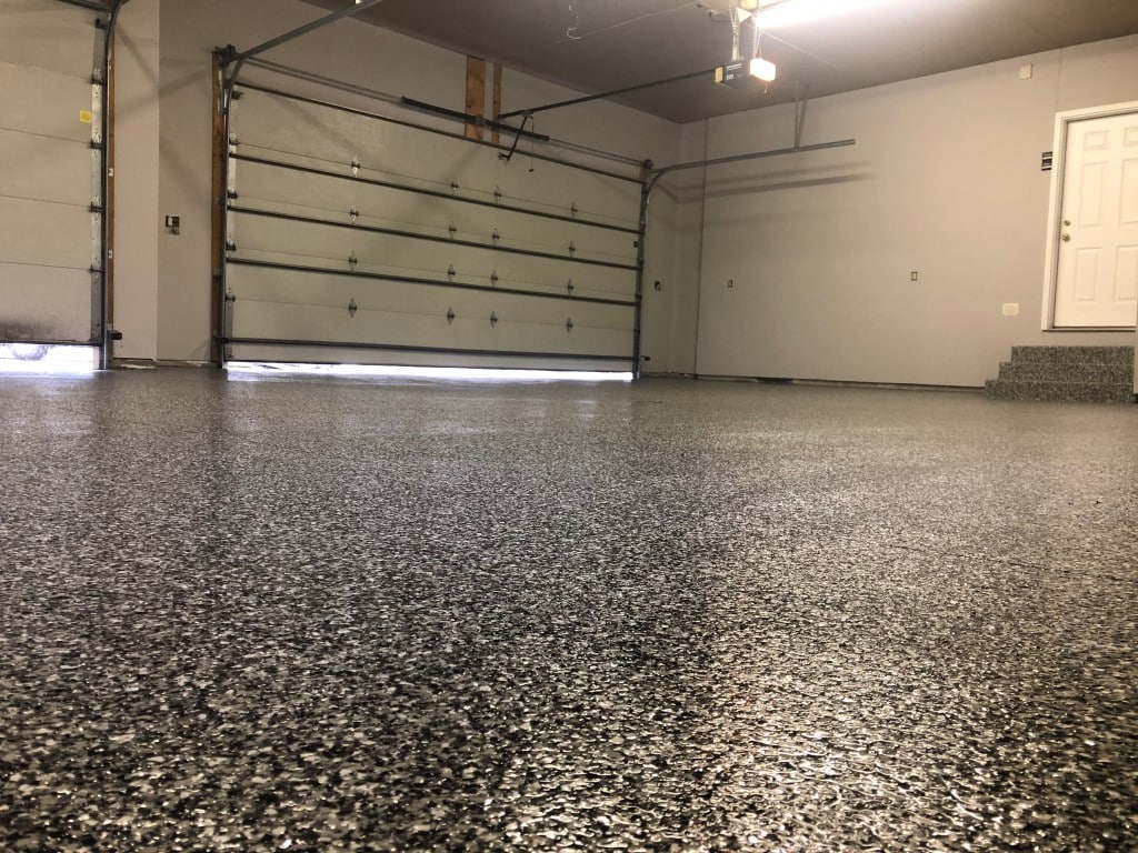 Clean garage with a polished, speckled floor, large closed garage door, beige walls, fluorescent lighting, and a small step leading to a side door.